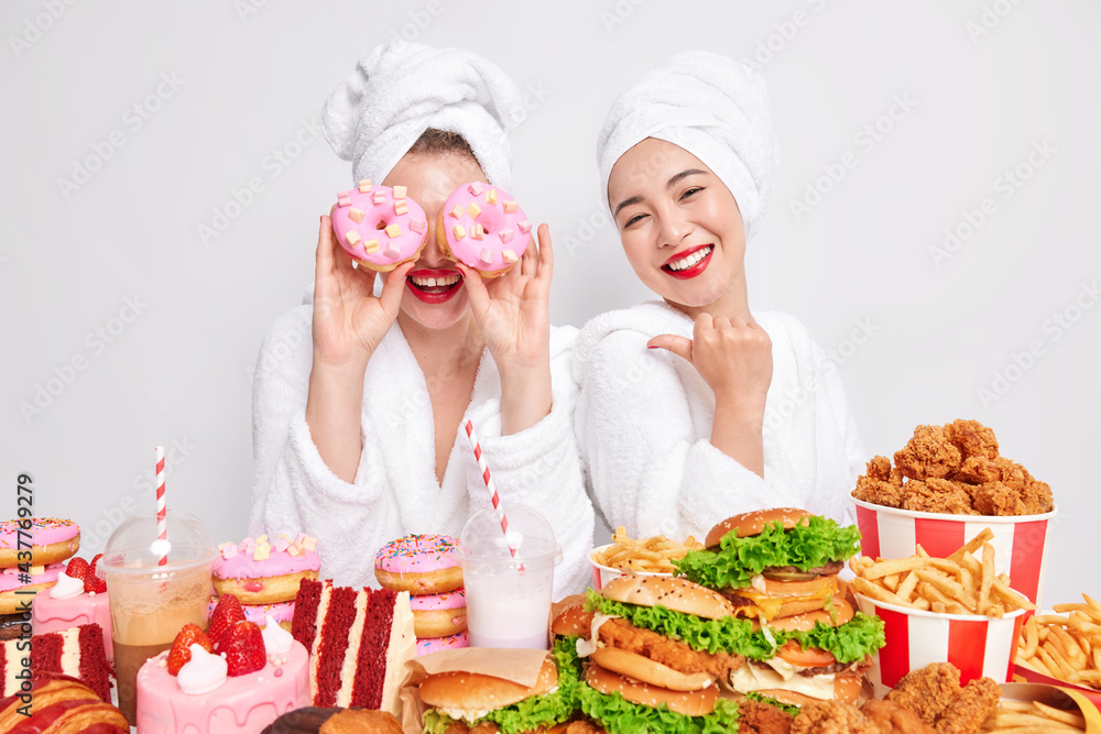 Positive woman points at her funny female friends who keeps delicious doughnuts over eyes as if goggles. Happy women enjoy eating junk food at home. Unhealthy nutrition and cheat meal concept