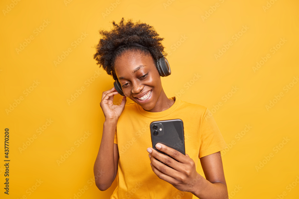 Studio shot of good looking dark skinned woman enjoys favorite playlist listens music via headphones uses smartphone dressed casually isolated over vivid yellow background. Technology concept Studio shot of good looking dark skinned woman enjoys favorite playlist listens music via headphones uses smartphone dressed casually isolated over vivid yellow background. Technology concept
