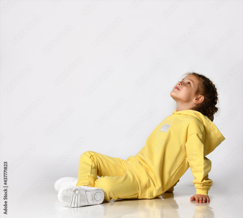 Stylish little girl sitting on a white background and dreamily looking up. Child with a beautiful hairstyle dressed in a yellow sports suit and sneakers looks towards the free space for text. Stylish little girl sitting on a white background and dreamily looking up. Child with a beautiful hairstyle dressed in a yellow sports suit and sneakers looks towards the free space for text.