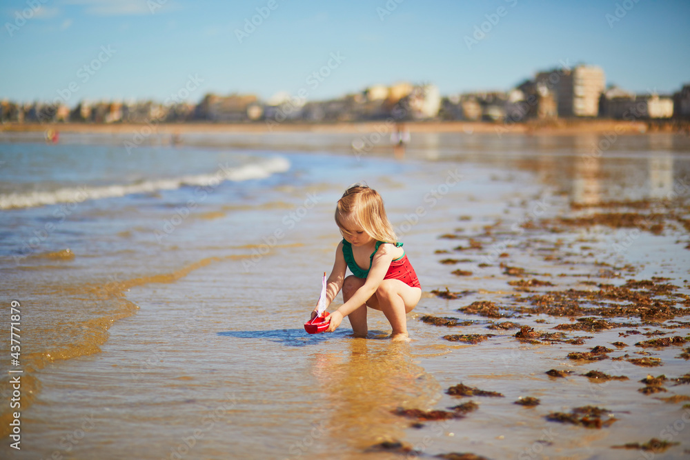 Fototapeta premium Adorable toddler girl in red swimsuit playing with wooden toy boat in water on the beach in Saint-Malo, Brittany, France