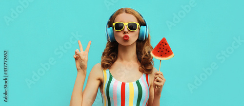 Summer fashion portrait of young woman in headphones listening to music with juicy slice of watermelon, female model blowing her lips posing on a colorful blue background