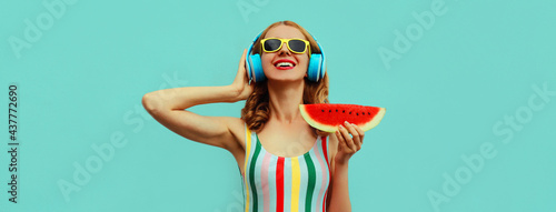 Summer portrait of cheerful happy smiling young woman in headphones listening to music with juicy slice of watermelon on a blue background