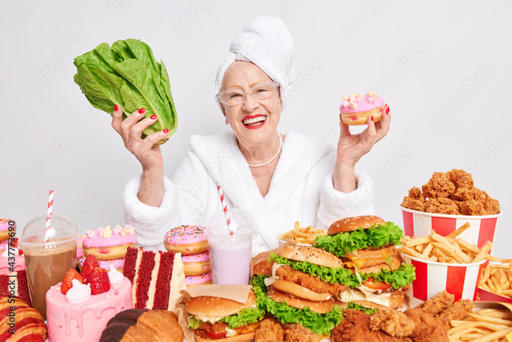 Positive aged European lady smiles happily wears domestic white bathrobe and towel wrapped over head holds delicious doughnut green vegerable chooses between junk food and healthy nutrition. Positive aged European lady smiles happily wears domestic white bathrobe and towel wrapped over head holds delicious doughnut green vegerable chooses between junk food and healthy nutrition.