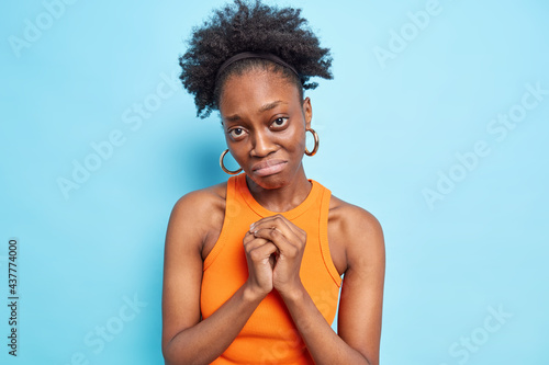 Disappointed dark skinned woman with guilty pity expression clasps hands asks for help or advice has some troubles pleads for apologize dressed in orange t shirt isolated over blue background photo