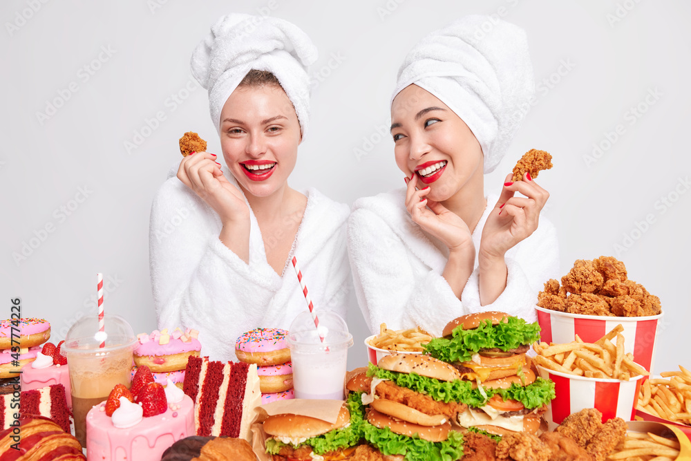 Happy mixed race young women look gladfully at each other eat junk food hold nuggets have tasty dinner dressed in bathrobes and towel on head. Overeating problem. Unhealthy nutrition concept Happy mixed race young women look gladfully at each other eat junk food hold nuggets have tasty dinner dressed in bathrobes and towel on head. Overeating problem. Unhealthy nutrition concept