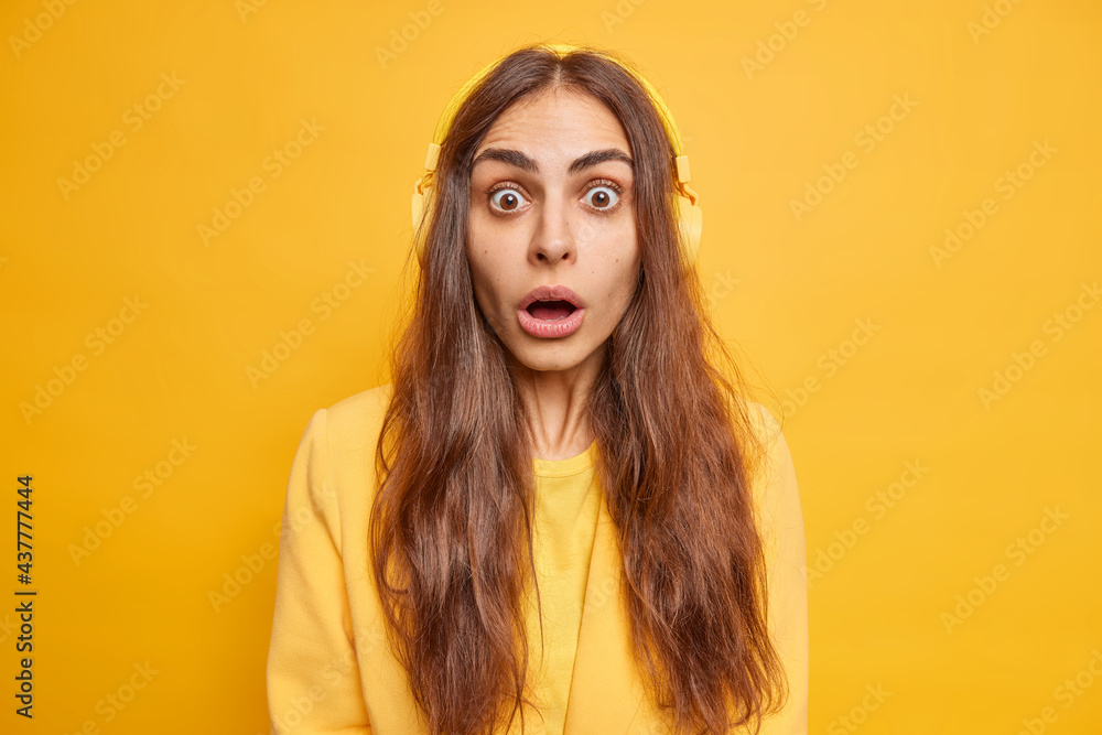 Shocked young European woman with long dark hair gasps surprised stands speechless keeps mouth opened listens radio via stereo headphones finds out amazing news isolated over yellow background Shocked young European woman with long dark hair gasps surprised stands speechless keeps mouth opened listens radio via stereo headphones finds out amazing news isolated over yellow background