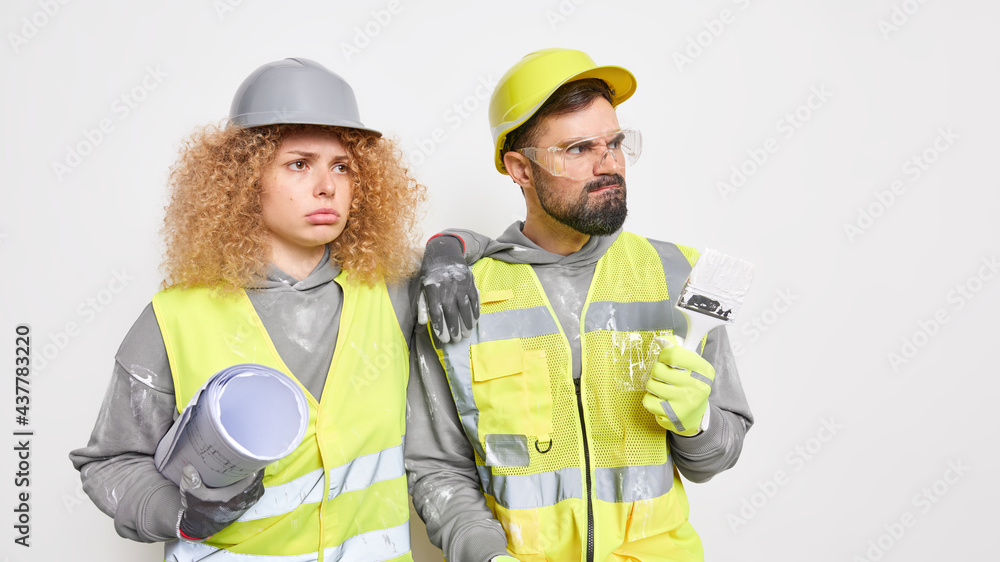 Home renovation and team work concept. Displeased woman architect man decorator work together on house redecoration pose with architectural blueprint painting brush isolated over white wall. Home renovation and team work concept. Displeased woman architect man decorator work together on house redecoration pose with architectural blueprint painting brush isolated over white wall.
