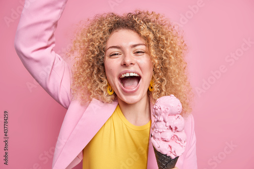 Studio shot of overjoyed curly haired woman has fun exclaims loudly from joy keeps arm raised eats tasty strawberry ice cream spends leisure day in merry company isolated over pink background