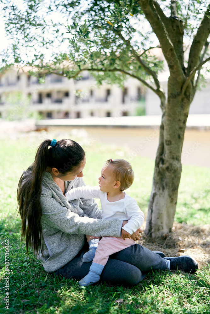 Mom holds a little girl in her arms, sits near a tree on the background of houses