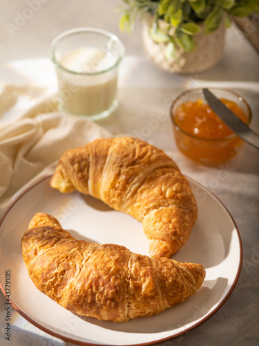 In the foreground of the photo  we see two croissants on a porcelain white plate. In the background  orange jam and milk in a glass glass. White silk tablecloth. Bright hues. Close-up.