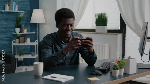 Cheerful happy black man playing video games on his phone in home office while taking a break from remote working from home. African american male having fun, enjoying enternainment on mobile phone photo