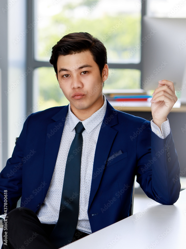 young attractive asian business man wearing navy blue suit with white shirt and necktie sitting down on the executive chair making confident face in office with natural lighting young attractive asian business man wearing navy blue suit with white shirt and necktie sitting down on the executive chair making confident face in office with natural lighting