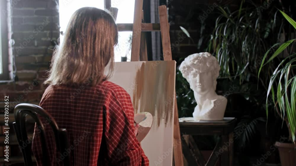 A young woman artist in a workshop applying base colors on a canvas with a small brush A young woman artist in a workshop applying base colors on a canvas with a small brush