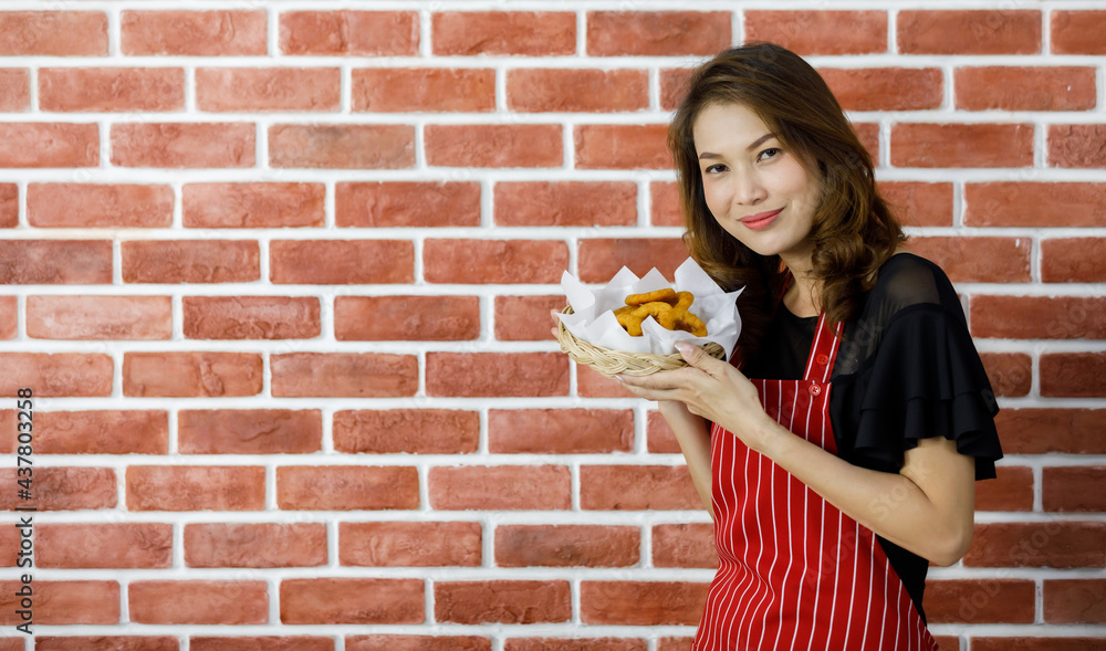 Attractive beautiful Asian woman in red striped apron standing near brick wall and smilingly persuade to enjoy eating favorite delicious food by raising fried snack and its tray to near mouth Attractive beautiful Asian woman in red striped apron standing near brick wall and smilingly persuade to enjoy eating favorite delicious food by raising fried snack and its tray to near mouth