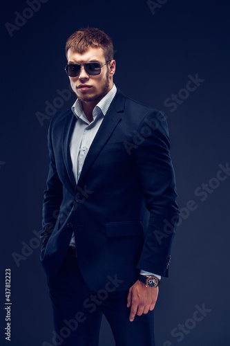 Fashion bearded young serious man in sunglasses, luxury suit looking at camera, one hand in pocket