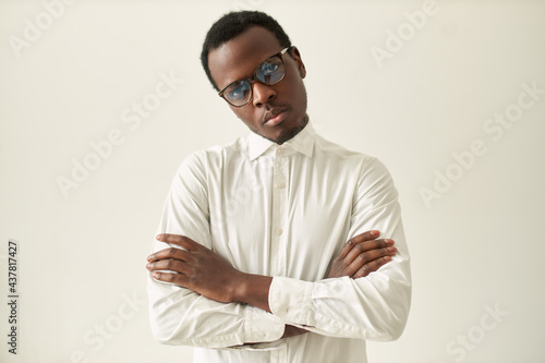 Horizontal shot of serious stubborn young black male in formal wear and eyeglasses keeping arms folded on his chest in closed posture, expressing disagreement or discontent. Body language