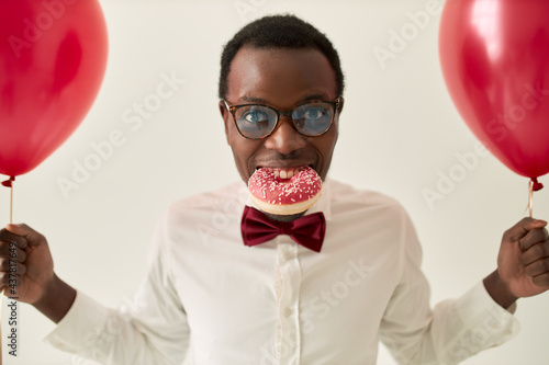 Portrait of funny young dark skinned man wearing elegant bow and glasses posing isolated with pink glazed doughnut in his mouth, holding red helium balloons, having fun at birthday party or wedding © Anatoliy Karlyuk