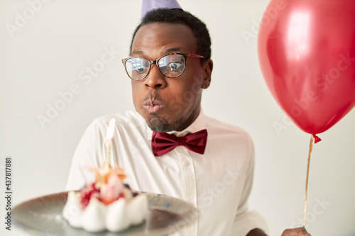 Portrait of funny young dark skinned male wearing eyeglasses and bow puffing cheeks, blowing out candle on birthday cake, trying hard, wishing his dreams will come true. Celebration and human emotions © Anatoliy Karlyuk
