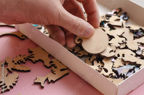 Open box with wooden jigsaw puzzle on pink background. A man's hand holds a fragment of a complex puzzle.