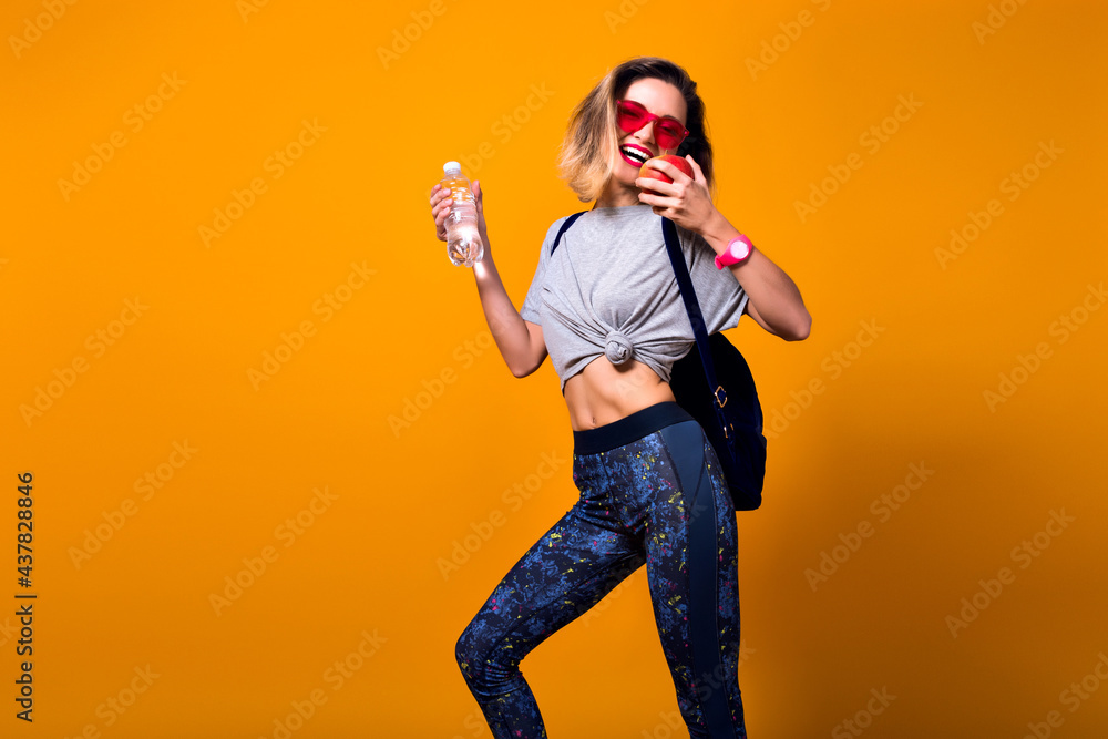 Slim girl with short hair with backpack going to gym and holding bottle of water. Laughing sporty young woman in sunglasses posing on bright background in studio with apple in hand. Slim girl with short hair with backpack going to gym and holding bottle of water. Laughing sporty young woman in sunglasses posing on bright background in studio with apple in hand.