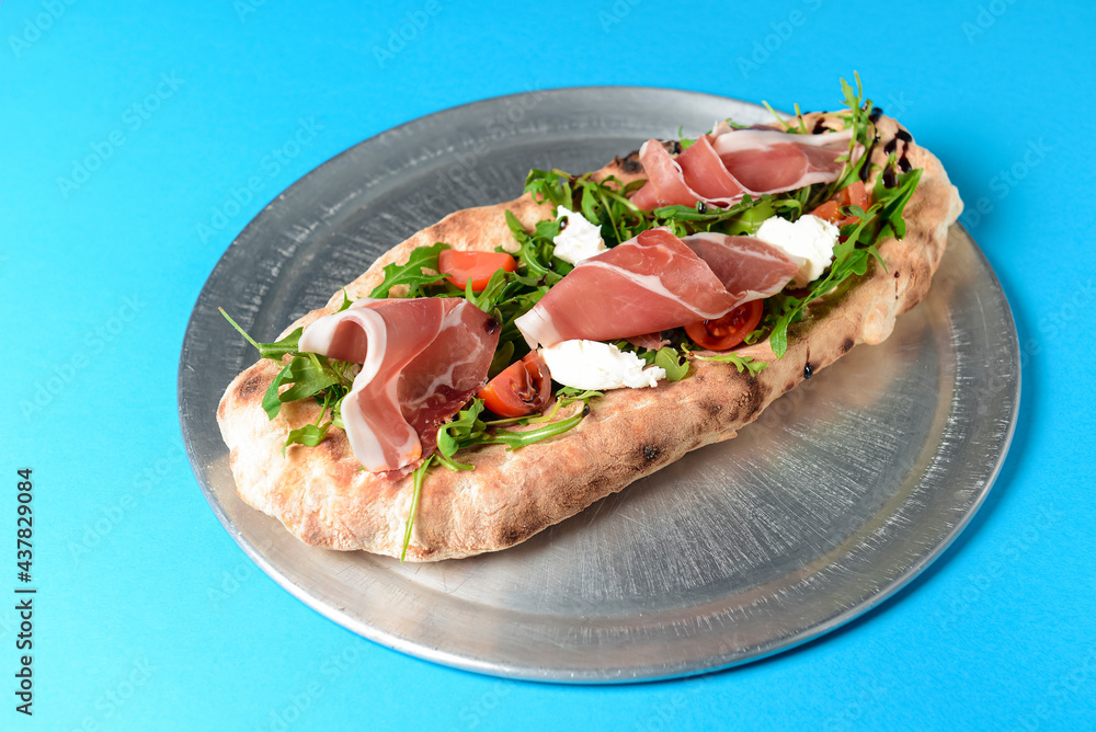 Sandwich with prosciutto, cheese and fresh arugula served on a metal tray over blue background.