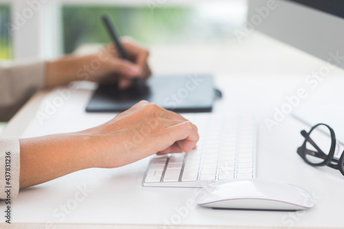 Close-up photo of designer's hand holding pen and working with graphics tablet in front of PC while working at home.