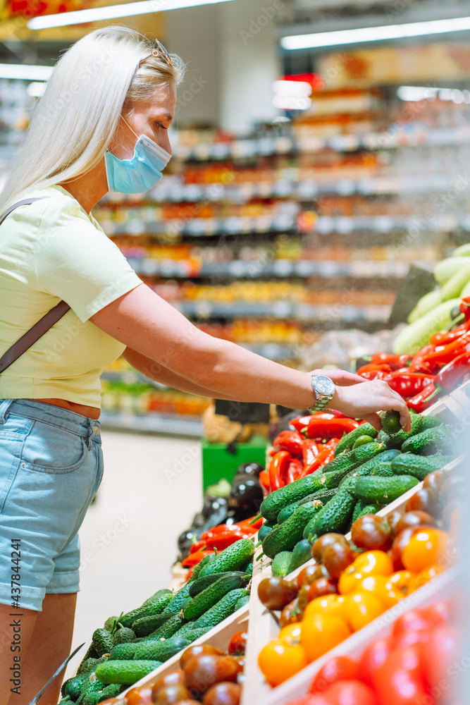 Young blonde woman wearing protective mask pick fresh vegetable on stall at market. Woman buying food Young blonde woman wearing protective mask pick fresh vegetable on stall at market. Woman buying food