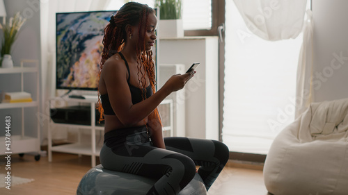 Slim woman with dark skin sitting on fitness ball writing message on phone during morning workout in living room. Fit adult in sportswear exercising abs muscle body while browsing online aerobic video