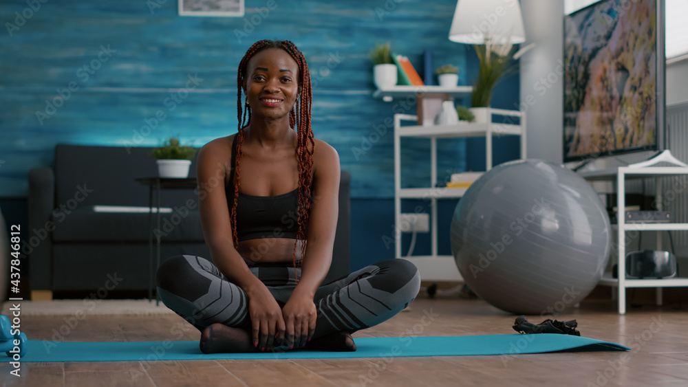 Portrait of black athlete sitting in lotus position on floor enjoying morning workout in living room. Woman in sportwear stretching muscle during strech gymnastics warming body Portrait of black athlete sitting in lotus position on floor enjoying morning workout in living room. Woman in sportwear stretching muscle during strech gymnastics warming body