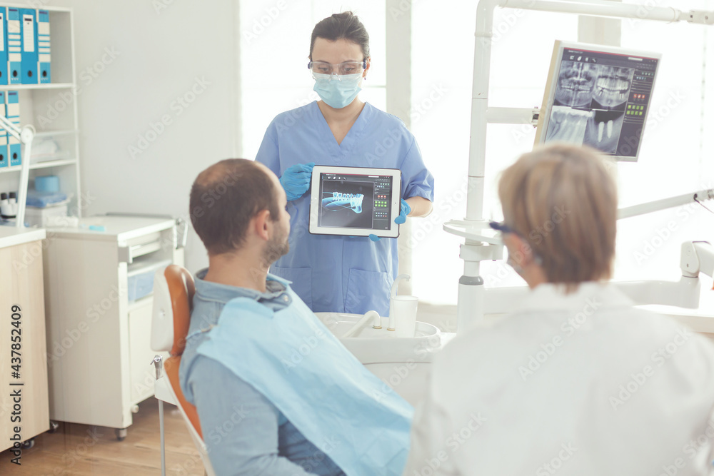 Stomatologist nurse pointing on digital screen explaining tooth x-ray to sick man sitting on dental chair in hospital stomatological office. Dentist doctor discussing toothache treatment Stomatologist nurse pointing on digital screen explaining tooth x-ray to sick man sitting on dental chair in hospital stomatological office. Dentist doctor discussing toothache treatment