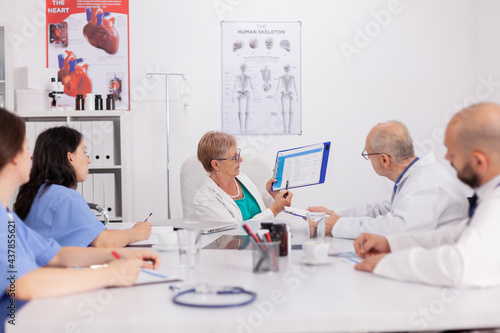 Senior pediatrician woman discussing sickness treatment using clipboard for medical presentation explaining disease expertise. Physiotherapist doctor discussing with hospital team in meeting room