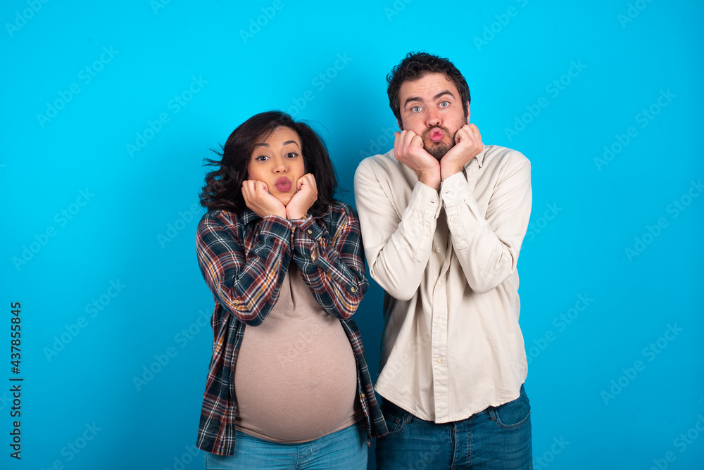 young couple expecting a baby standing against blue background with surprised expression keeps hands under chin keeps lips folded makes funny grimace young couple expecting a baby standing against blue background with surprised expression keeps hands under chin keeps lips folded makes funny grimace