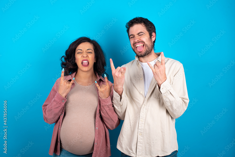 young couple expecting a baby standing against blue background making rock hand gesture and showing tongue young couple expecting a baby standing against blue background making rock hand gesture and showing tongue