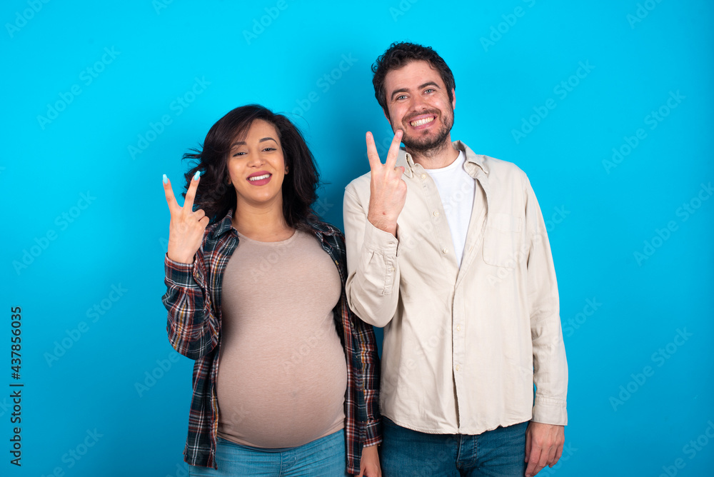 young couple expecting a baby standing against blue background smiling and looking friendly, showing number two or second with hand forward, counting down young couple expecting a baby standing against blue background smiling and looking friendly, showing number two or second with hand forward, counting down