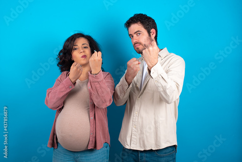 young couple expecting a baby standing against blue background Ready to fight with fist defense gesture, angry and upset face, afraid of problem.
