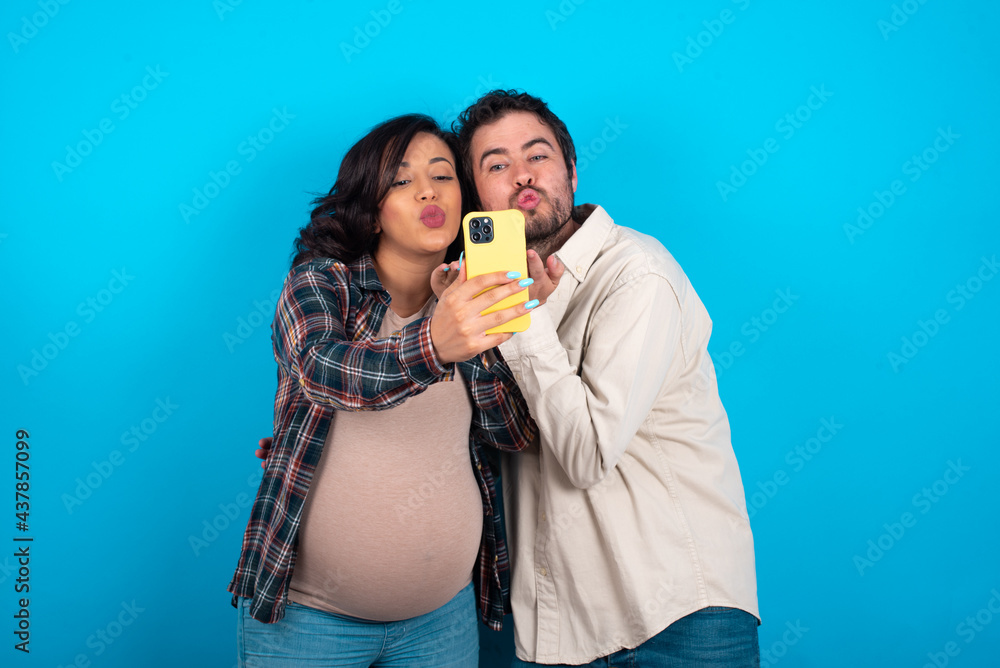 young couple expecting a baby standing against blue background blows air kiss at camera of smartphone and takes selfie, sends mwah via online call. young couple expecting a baby standing against blue background blows air kiss at camera of smartphone and takes selfie, sends mwah via online call.