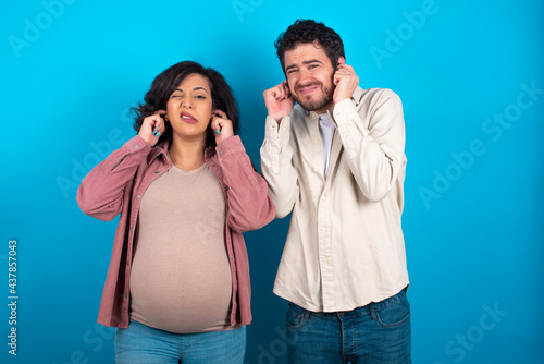 young couple expecting a baby standing against blue background covering ears with fingers with annoyed expression for the noise of loud music. Deaf concept.