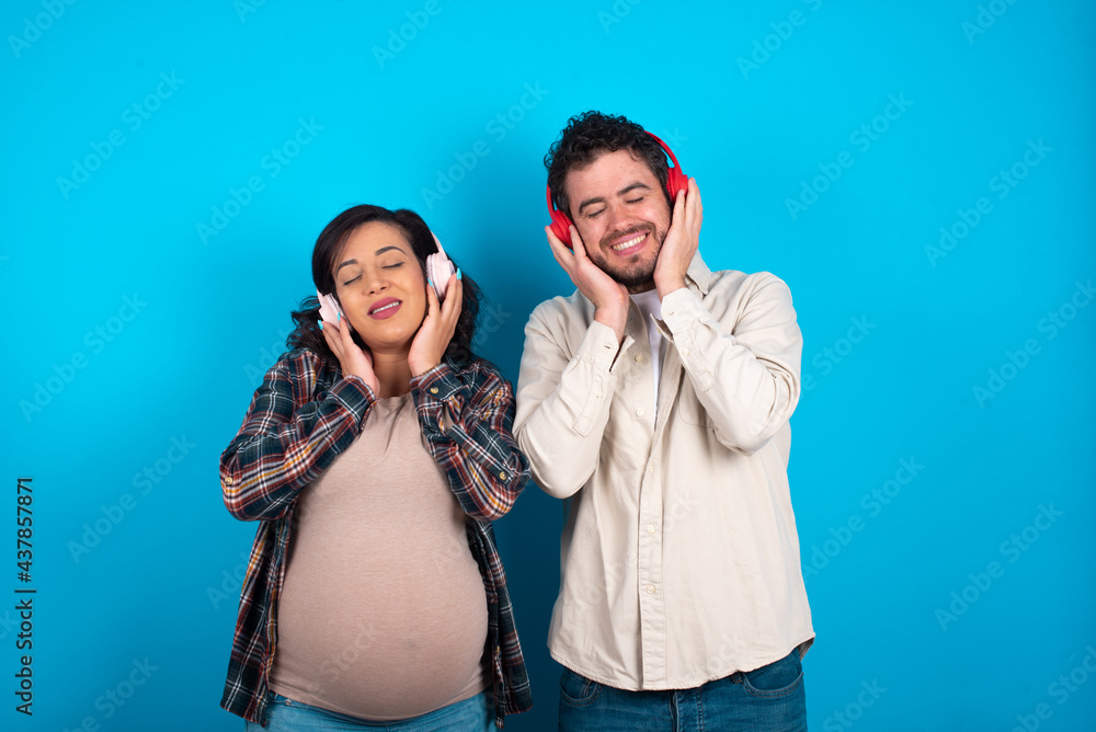 Pleased young couple expecting a baby standing against blue background enjoys listening pleasant melody keeps hands on stereo headphones closes eyes. Spending free time with music Pleased young couple expecting a baby standing against blue background enjoys listening pleasant melody keeps hands on stereo headphones closes eyes. Spending free time with music