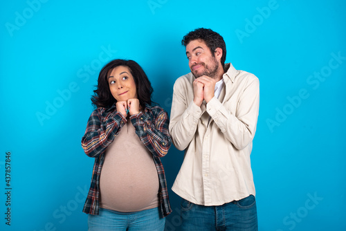 Curious young couple expecting a baby standing against blue background keeps hands under chin bites lips and looks with interest aside.