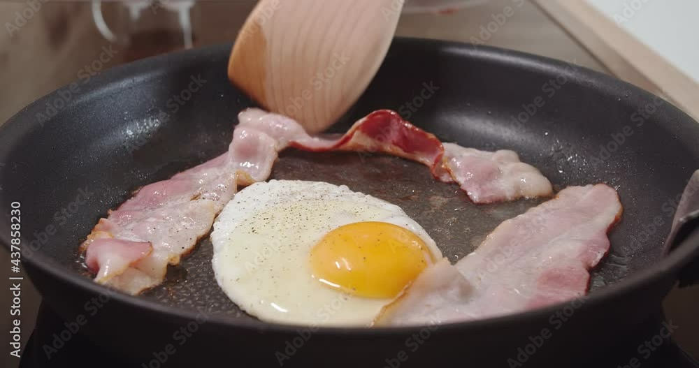 Egg and bacon slices are fried in a hot skillet. Spices are visible on them. Someone gently straightens food with a spatula so that it is evenly fried Egg and bacon slices are fried in a hot skillet. Spices are visible on them. Someone gently straightens food with a spatula so that it is evenly fried