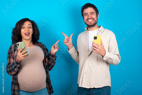 Astonished young couple expecting a baby standing against blue background holding her telephone and pointing with finger aside at empty copy space