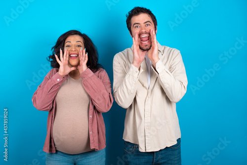 young couple expecting a baby standing against blue background shouting excited to front.