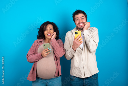 Joyous young couple expecting a baby poses with mobile phone device, types text message on modern smartphone, watches funny video during free time, enjoys good internet connection,