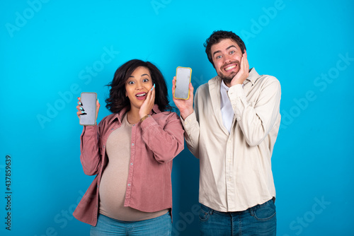 young couple expecting a baby standing against blue background hold hand modern technology use touch face palm astonished impressed scream wow omg unbelievable unexpected