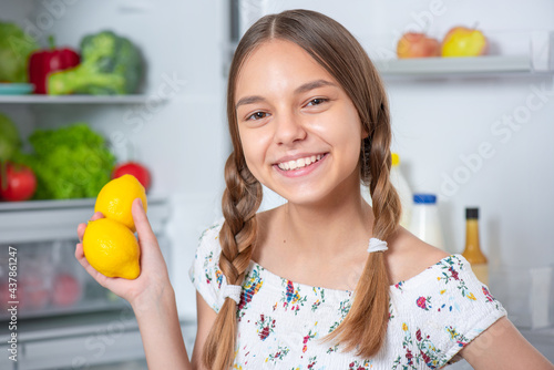 Beautiful young happy teen girl holds fresh yellow lemons while standing near open fridge in kitchen at home