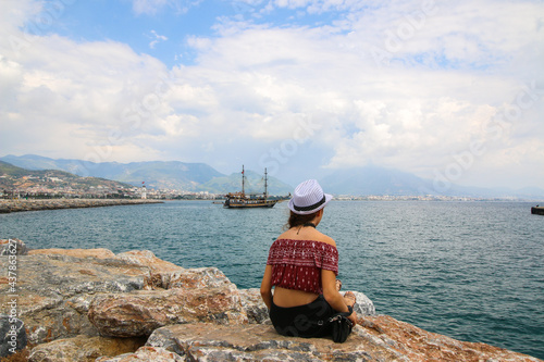 A single girl with white hat sits on a rock on the shore against the sky and the mediterranean sea, lifestyle, recreation, solitude, meditation, copy space.