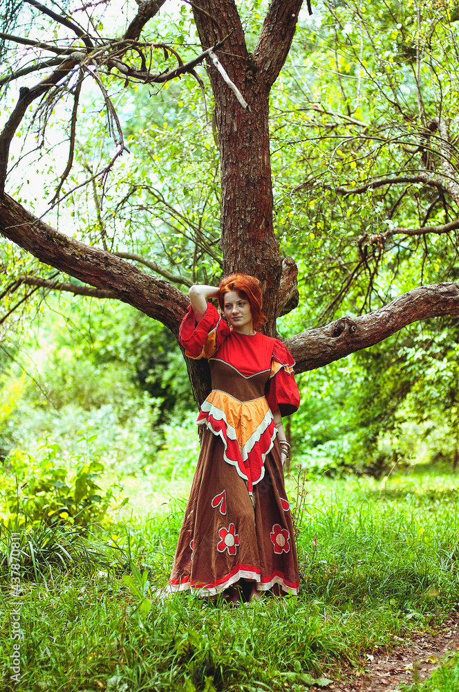 Stylish girl with freckles and red hair in red long dress dancing at green wild forest on nature. Portrait of boho woman posing in summer countryside. Happy lifestyle. Atmospheric moment. 