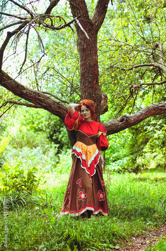 Stylish girl with freckles and red hair in red long dress dancing at green wild forest on nature. Portrait of boho woman posing in summer countryside. Happy lifestyle. Atmospheric moment. 