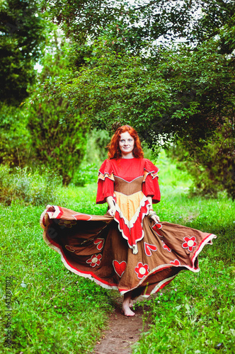 Stylish girl with freckles and red hair in red long dress dancing at green wild forest on nature. Portrait of boho woman posing in summer countryside. Happy lifestyle. Atmospheric moment. 