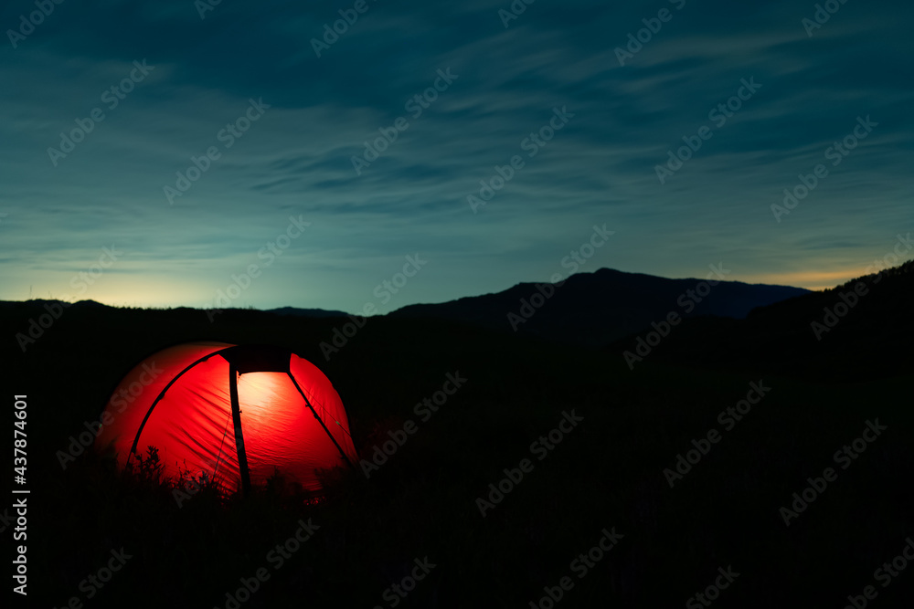 Glowing red tent with hikers sleeping in nature under dramatic night sky with noctilucent clouds. Lighted camping tent located in a wild area during the summer. Copy space.